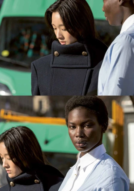 Two women in a fashion shot. An Asian woman wears a dark coat with brass buttons. An African woman wears a light blue shirt.