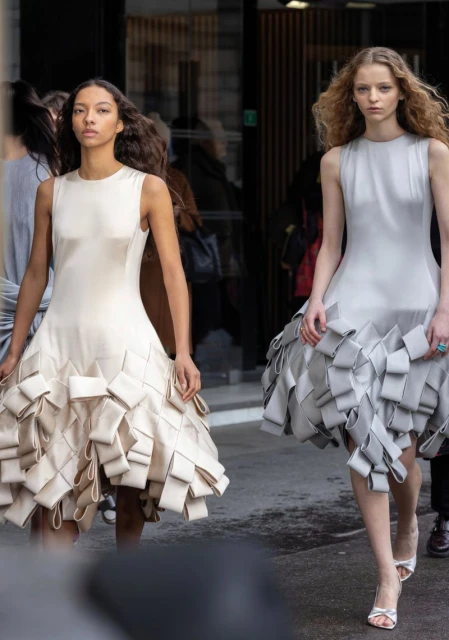 Two fashion models in sleeveless dresses with unique ribbon detail walk a runway during an outdoor fashion show on a cloudy day.