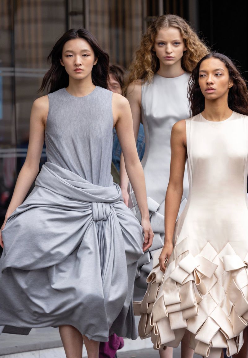 Three fashion models walk during a runway show, wearing unique grey and ivory sleeveless dresses with draped and folded design elements.