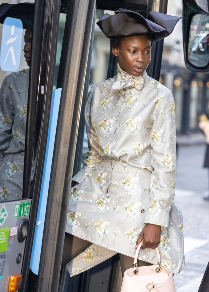 Stylish adult female in floral print shirt dress, dark tights, hat, and purse, standing on a bus with gray and blue accents.