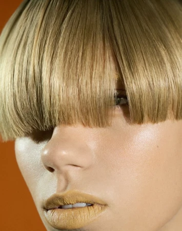 Close-up portrait of a woman with blonde bowl-cut hairstyle, wearing gold lipstick, set against a vivid orange background in a beauty shot.