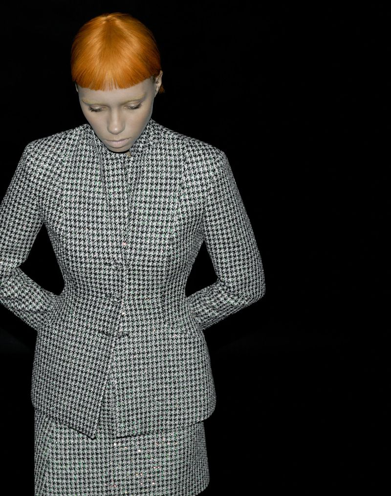 Model with bright orange hair wearing a houndstooth jacket and matching skirt against a solid black background.