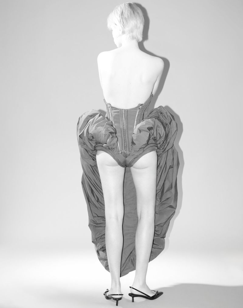 Back view of adult woman with short blonde hair wearing corset and skirt with high heel sandals in a photo studio setting.