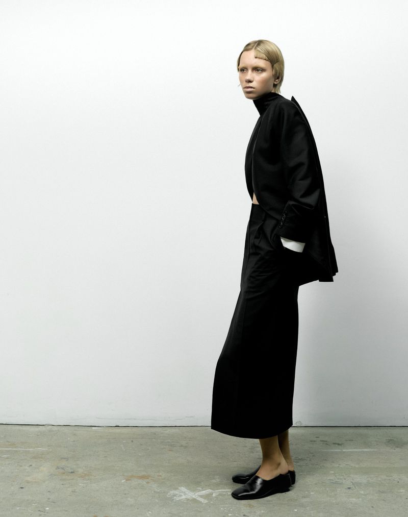 Side profile shot of a single young adult female in a black outfit against a white background in a studio, minimalist fashion photograph.