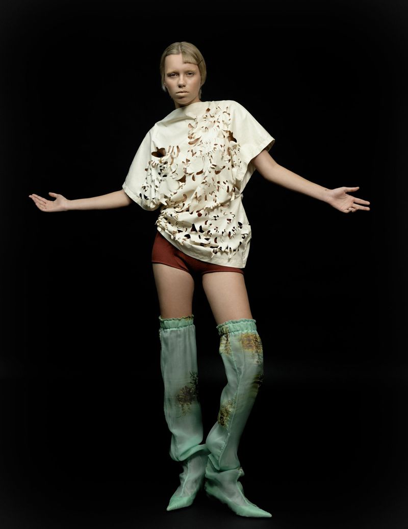 Fashion shot of a female adult model in a cut-out patterned top and thigh-high socks, standing against a black backdrop in a studio.