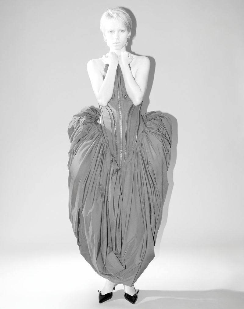 Monochrome fashion shot of a woman with short blonde hair wearing a dramatic, draped couture dress and black heels against a plain backdrop.
