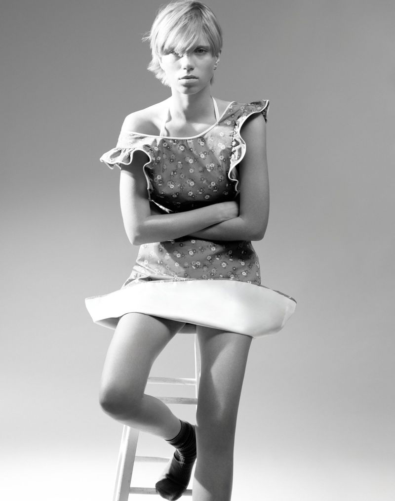 Model with short hair seated on a stool wearing a patterned dress against a grey studio backdrop