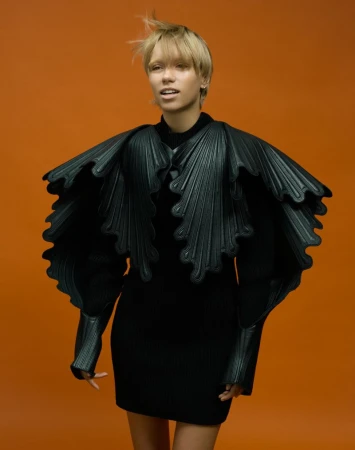 Model Lena Mantler in black sculptural dress and pleated cape against solid orange backdrop for 10 Magazine FACE VALUE.