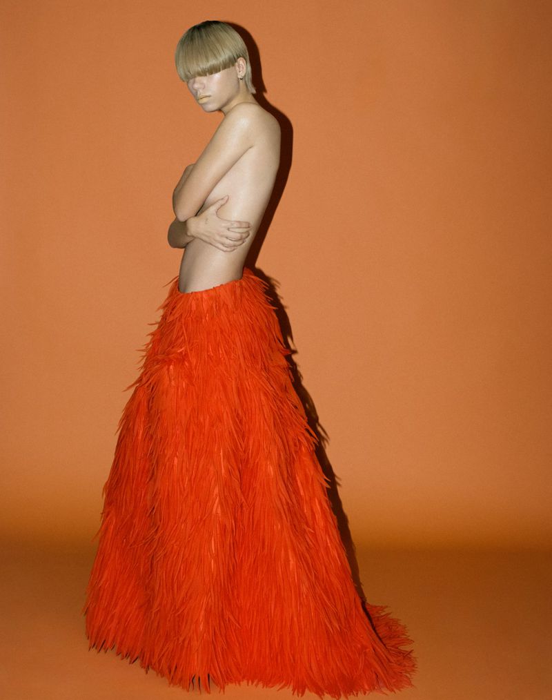 Androgynous individual poses in studio wearing orange feathered skirt, arms crossed, gazing at camera against orange background.