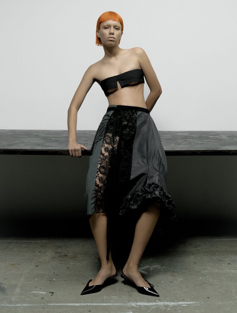 Fashion shot of a woman with bright orange hair wearing a black bandeau top and dark skirt with lace, standing indoors on a concrete surface.