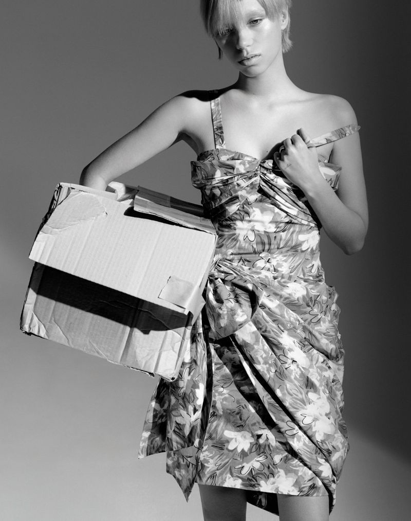 Monochrome fashion portrait of a woman in floral dress holding a cardboard box in a neutral studio setting.