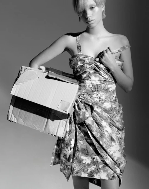 Monochrome fashion portrait of a woman in floral dress holding a cardboard box in a neutral studio setting.