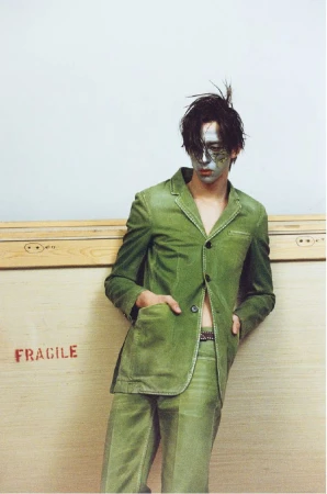 A person wearing a green suit and silver face makeup leans against a wooden crate with the word 'fragile' printed on it.