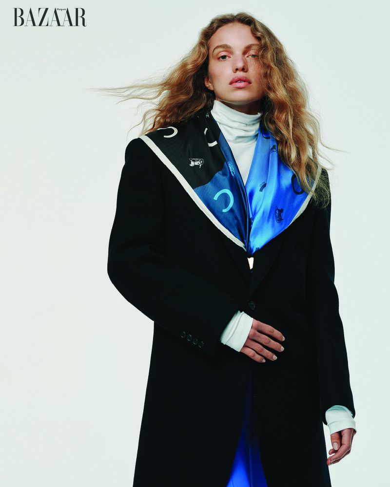 Fashion editorial shot featuring a woman with curly hair wearing a black coat with a white turtleneck and a blue-and-black patterned scarf.