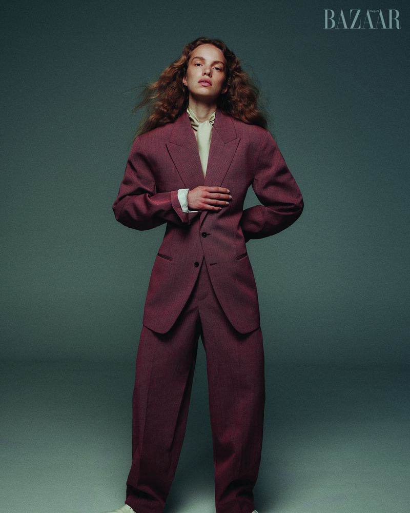 Fashion editorial shot of a red-haired woman wearing an oversized red suit, posing in a studio with diffused lighting, and a BAZAAR logo.