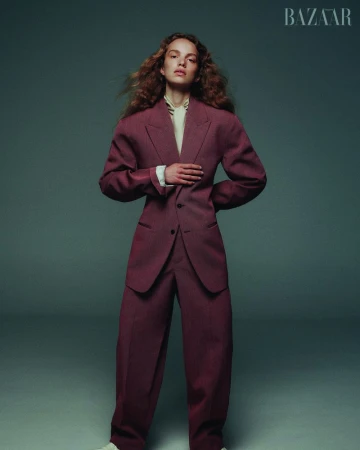 Fashion editorial shot of a red-haired woman wearing an oversized red suit, posing in a studio with diffused lighting, and a BAZAAR logo.