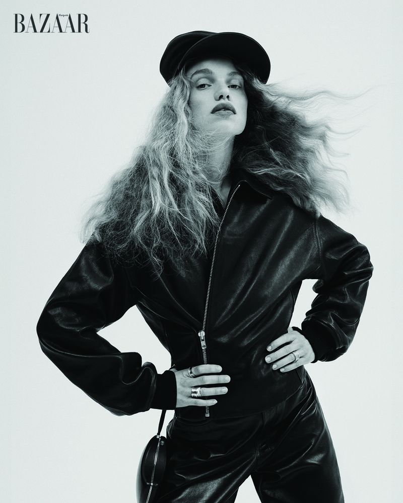 Fashion shot of woman with long wavy hair in a black leather jacket and cap, hands on waist, posing in front of a white background.