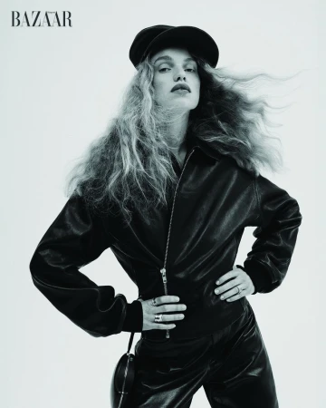 Fashion shot of woman with long wavy hair in a black leather jacket and cap, hands on waist, posing in front of a white background.