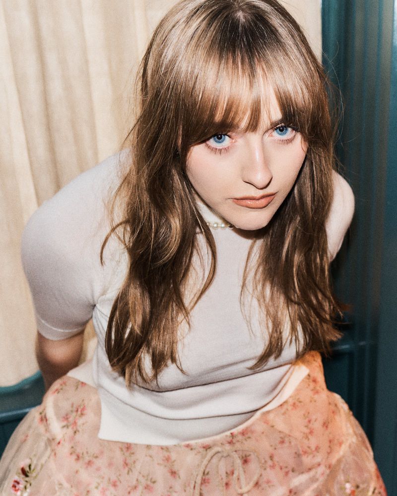 Portrait of a young woman with blue eyes and brown hair, wearing a white top and floral skirt, posing indoors with natural lighting.