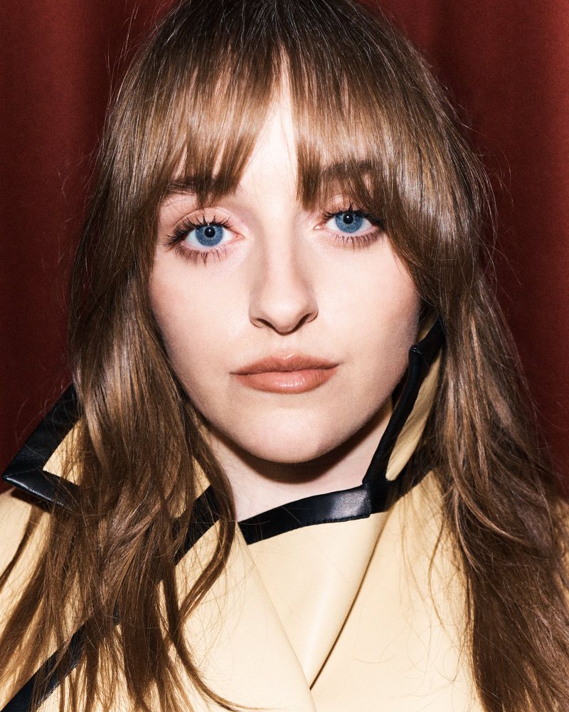 Close-up portrait of a young adult woman with blue eyes and brown hair wearing a yellow coat in front of a red background.