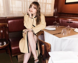 Musician Nell Mescal seated at a restaurant booth wearing a pale yellow coat with black piping for FT HTSI Magazine.