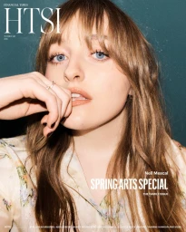 Close-up portrait of musician Nell Mescal with long brown hair and blue eyes, wearing a patterned top against a dark green textured background.