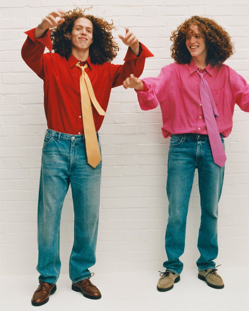 Two adult males pose indoors against a white brick wall, wearing colorful shirts and ties with blue jeans and dress shoes.