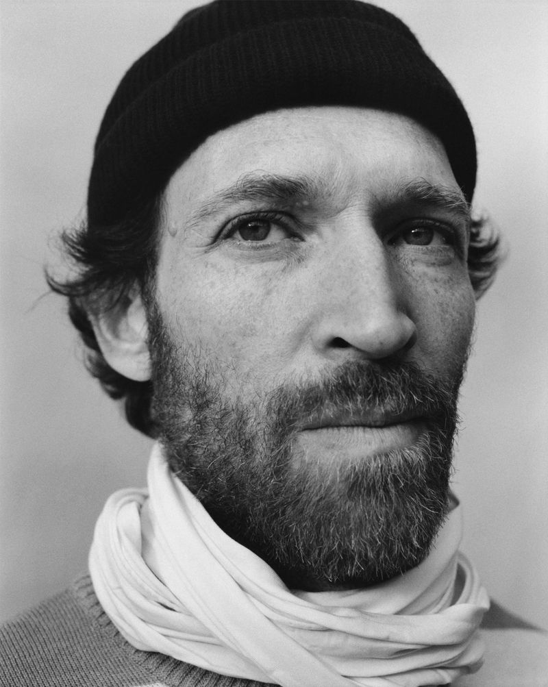 Black and white portrait of adult male wearing a beanie and scarf, with a beard, in a close-up shot in front of a plain backdrop.