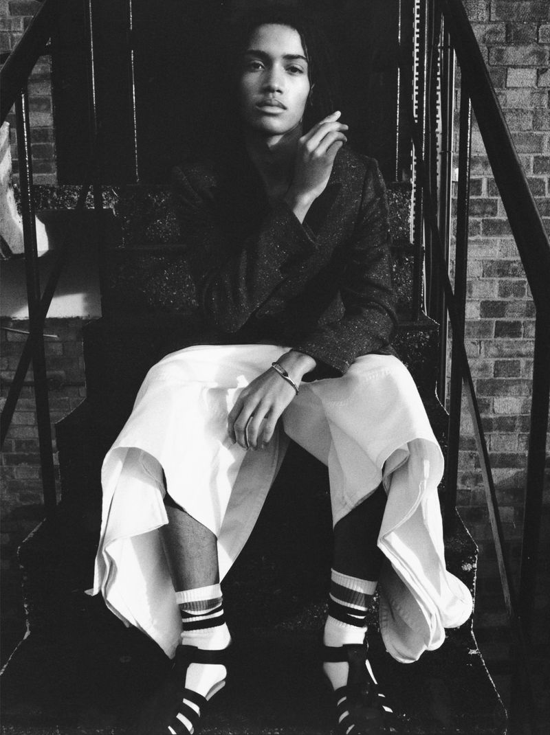 Black and white portrait of a woman sitting on metal stairs in a tweed jacket and white culottes, wearing striped socks and sandals.