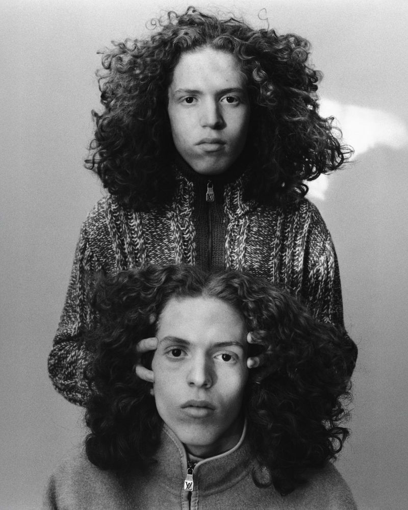 Two young adult males pose in a black and white portrait; the standing figure in a cardigan, the seated figure with hands near his head.