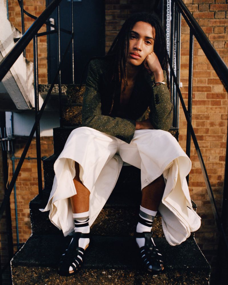 Adult male with dreadlocks wearing a green jacket and white pants, sitting on gray stairs with brick building in the background.