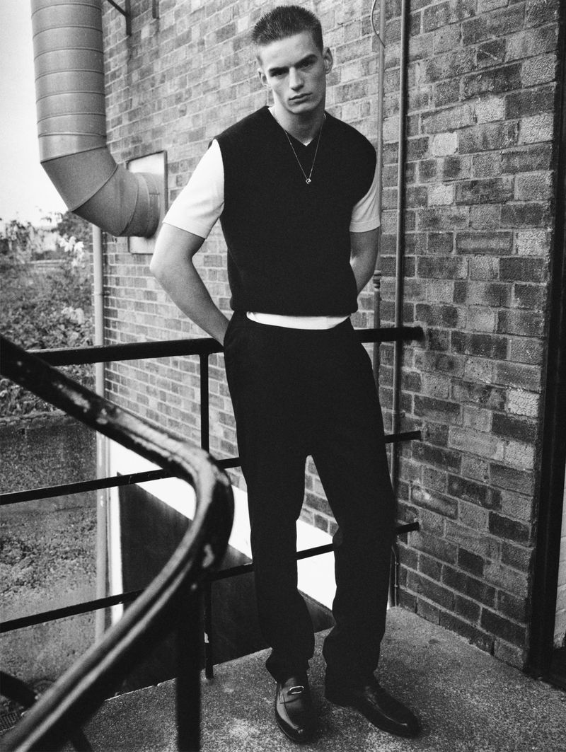 Black and white full shot of a young adult male posing in front of a brick wall wearing a sweater vest and dress pants.