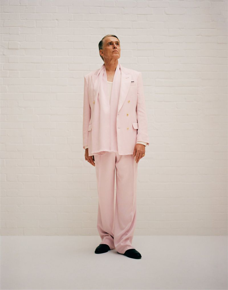 Full body shot of an adult male in a pale pink suit and scarf, standing against a white brick wall, looking upwards.
