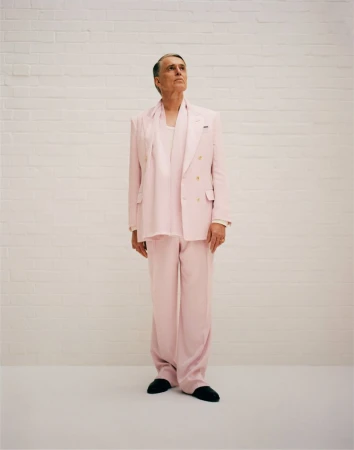 Full body shot of an adult male in a pale pink suit and scarf, standing against a white brick wall, looking upwards.