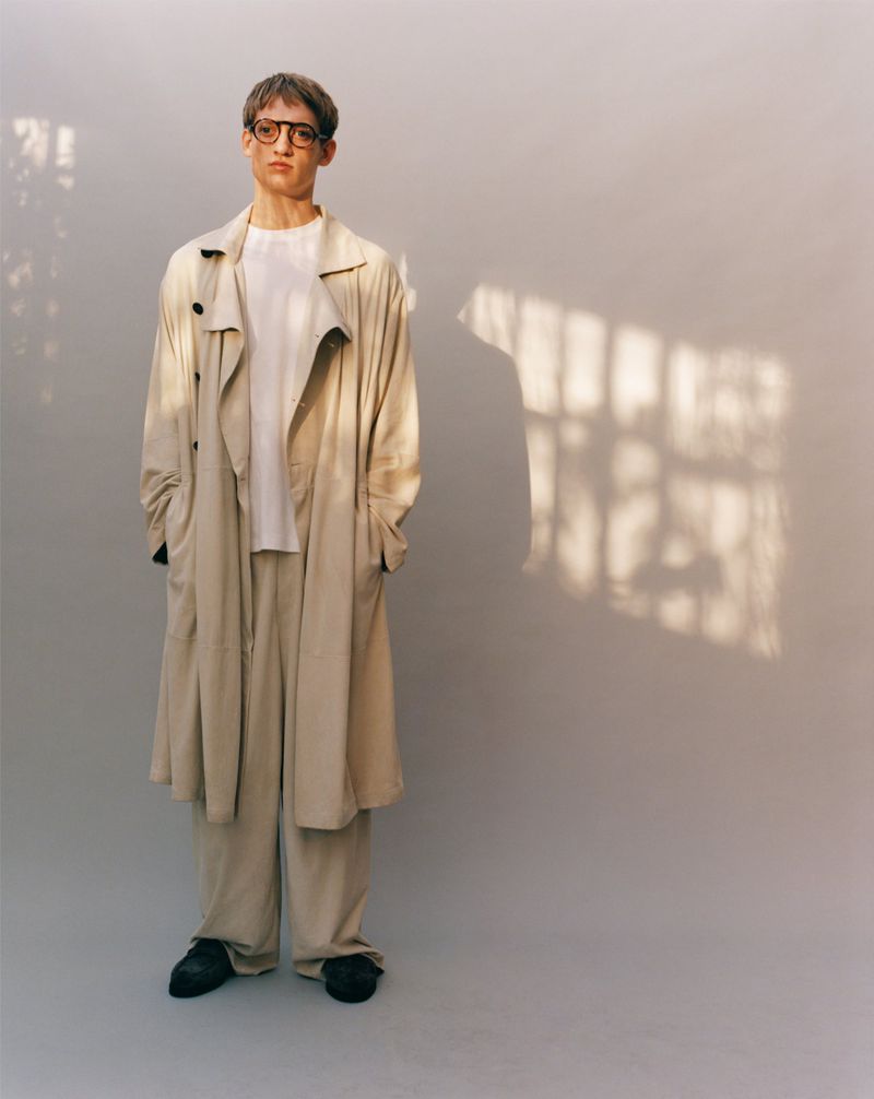 Man in glasses and beige trench coat stands in a light-filled studio with long sleeves and relaxed fit pants in a fashion pose.