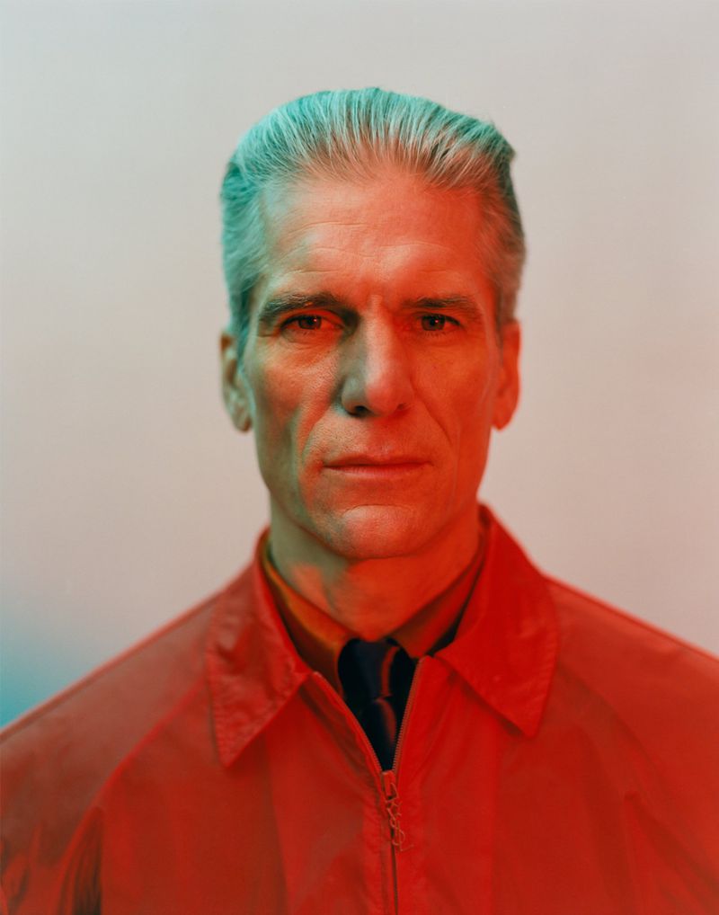 Headshot of an older adult male with grey hair wearing a red jacket, set against a gradient background with red and blue lighting.