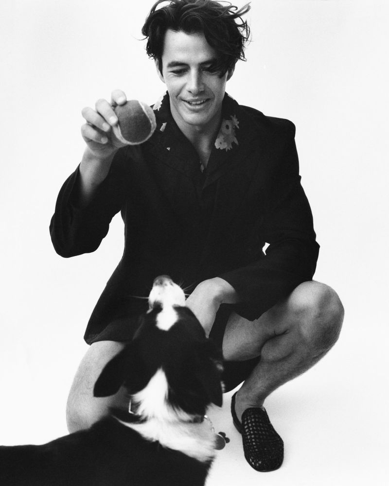 Monochrome portrait of a man squatting with a tennis ball, looking at a dog reaching up, set against a plain white background.