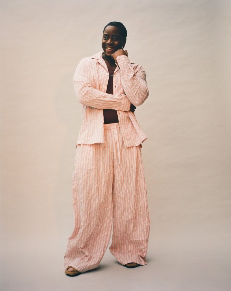 Full body shot of smiling adult male in a pink striped linen shirt and pants, standing in front of a neutral colored background.