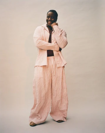 Full body shot of smiling adult male in a pink striped linen shirt and pants, standing in front of a neutral colored background.