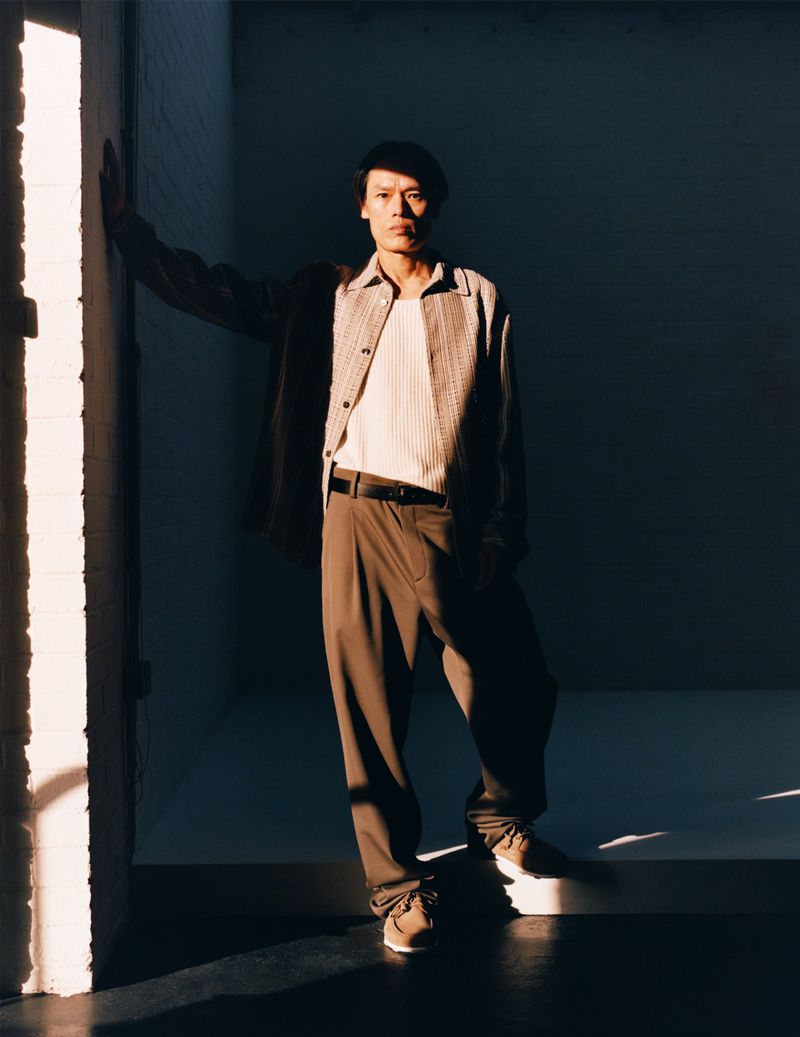 Portrait of an adult male in casual attire standing indoors. He wears a jacket, pants, and sneakers, with sunlight casting shadows on the wall.