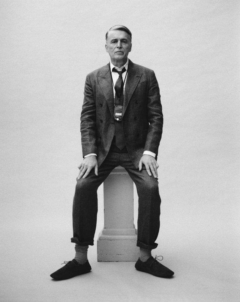 Black and white portrait of an adult man in a suit sitting on a pedestal in a photo studio with a plain white background.