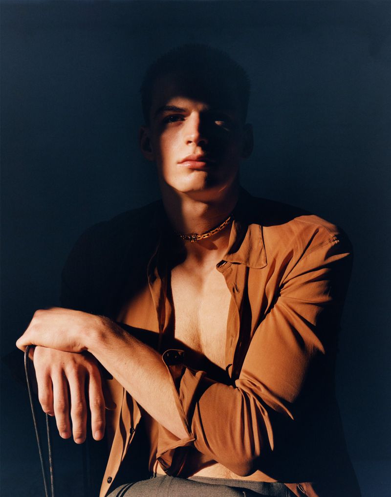 Portrait of a male model with short hair wearing a brown shirt and gold chain, sitting in a partially shadowed environment.