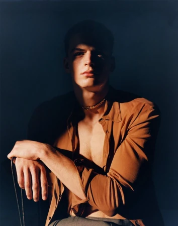 Portrait of a male model with short hair wearing a brown shirt and gold chain, sitting in a partially shadowed environment.