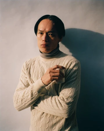 Portrait of a serious-looking Asian man wearing a white cable-knit sweater, holding a cup indoors against a white wall with soft shadows.