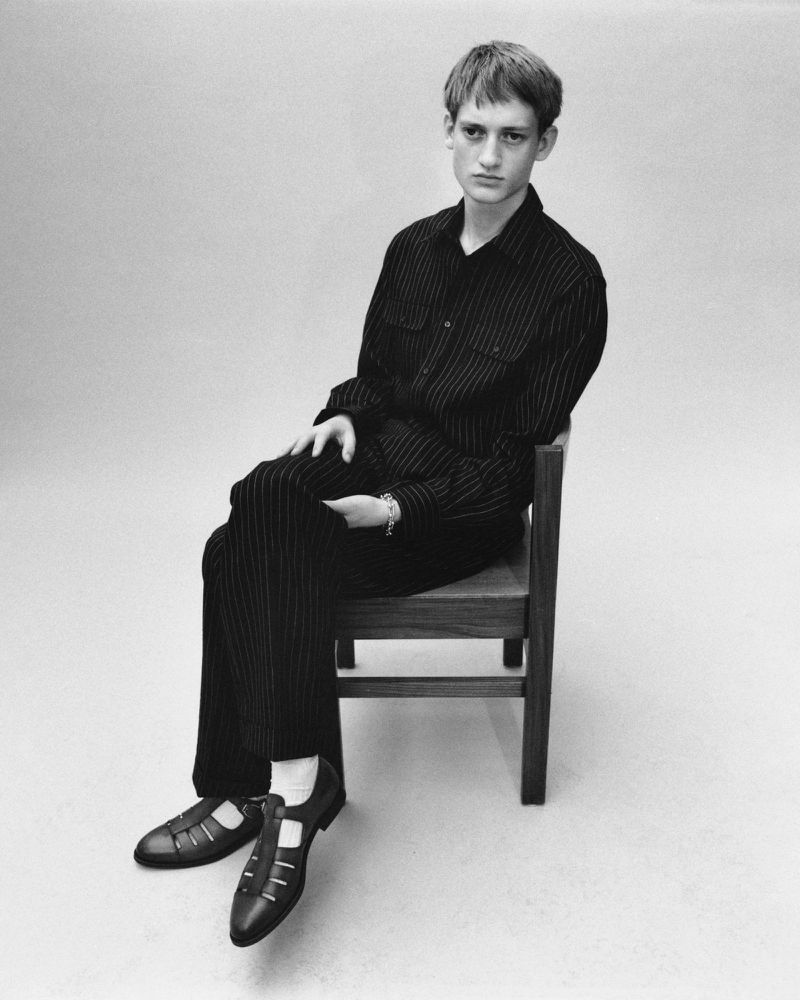 Black and white portrait of a young adult male seated on a wooden chair in a studio wearing a pinstripe jumpsuit and leather shoes.