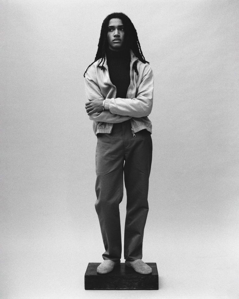 Full-length studio portrait of a Black man with dreadlocks wearing a jacket, turtleneck, and pants, standing on a box against a solid backdrop.