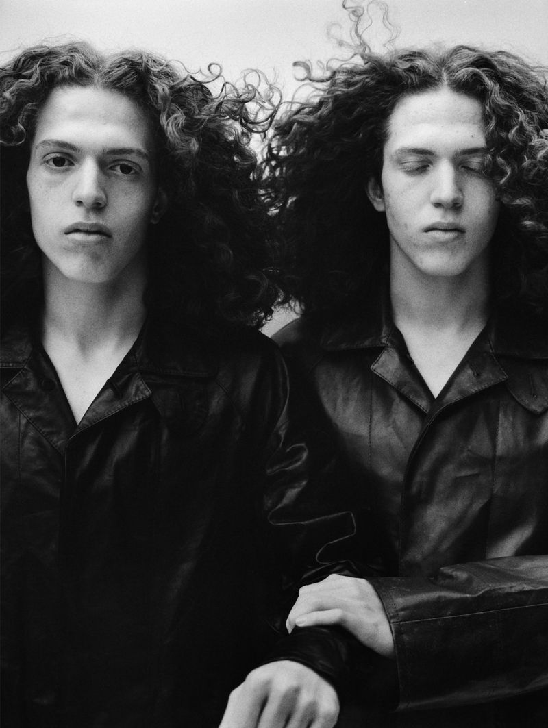 Black and white portrait of two young adult men with curly dark hair, wearing matching black collared jackets, and light skin.