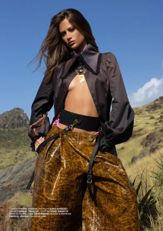 Fashionable woman poses outdoors against blue sky wearing brown top, wide belt, and shiny tan trousers in a fashion editorial shot.