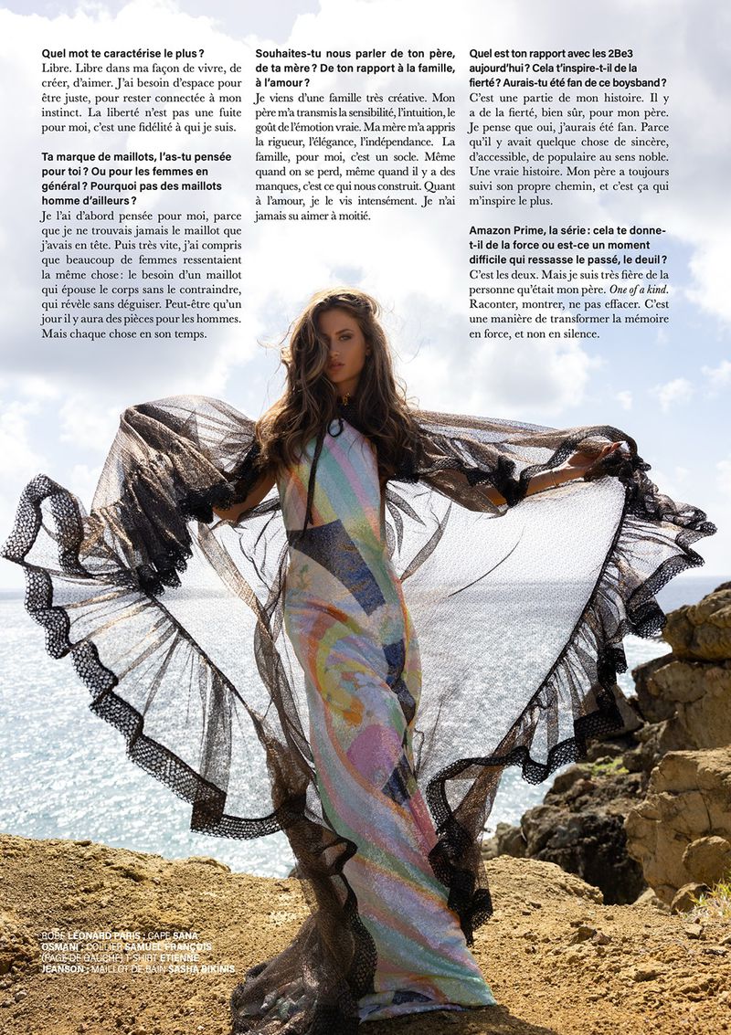 A woman poses on a rocky outcrop with the ocean in the background, wearing a flowing, colorful dress and sheer black cape. 