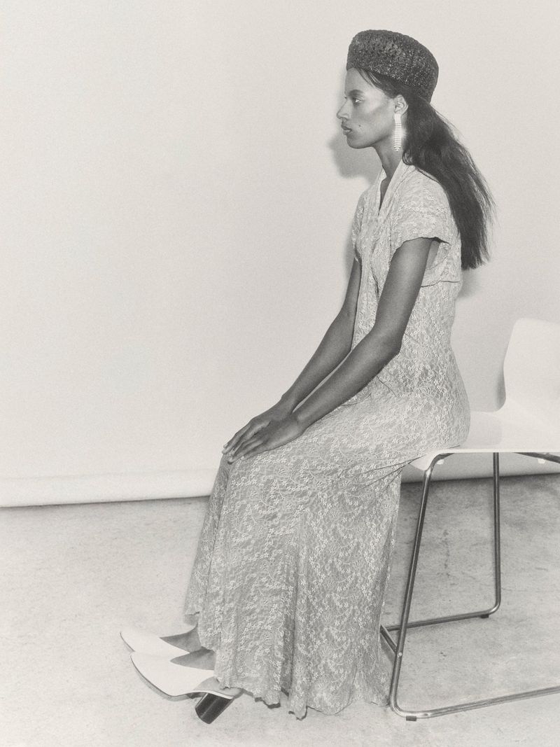Woman in a patterned dress and hat seated on a white chair in a photo studio with neutral lighting and backdrop.